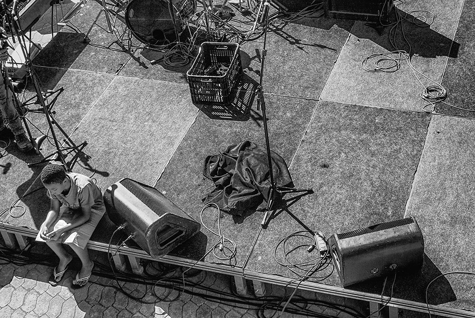 Fotografia em preto e branco de um menino sentado em uma estrutura elevada de concreto (palco), olhando para baixo em direção a uma área urbana com equipamentos de som e grades em Belo Horizonte.