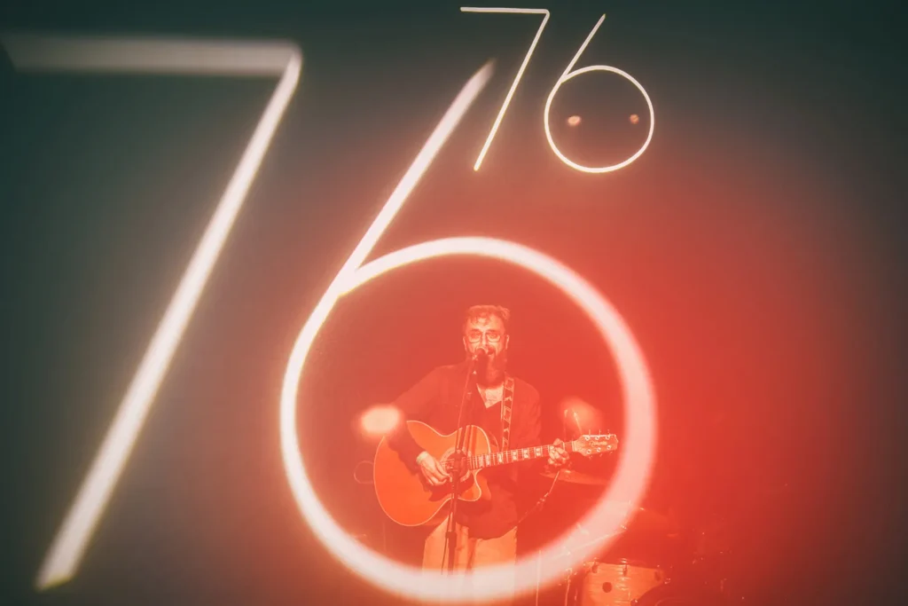 Fotografia em dupla exposição de Ricardo Laf mostrando o músico Péricles Garcia no palco, envolto por um círculo de luz que forma o número 76.