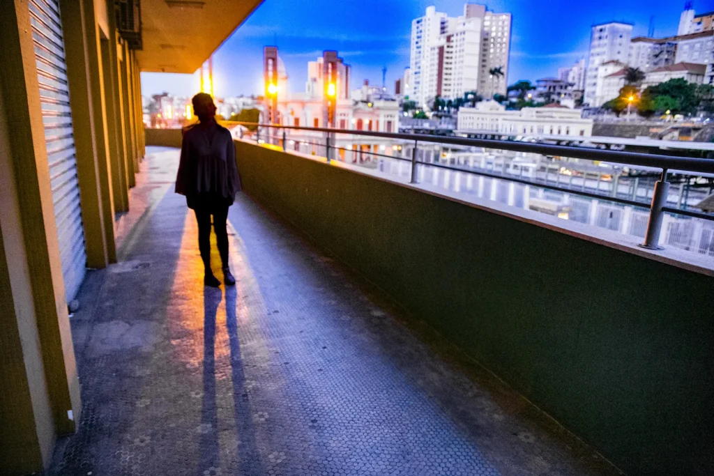 Fotografia colorida de Ricardo Laf mostrando a silhueta escura de uma pessoa de costas, observando a paisagem urbana do Baixo Centro de Belo Horizonte a partir de uma estrutura elevada com guarda-corpo metálico durante o crepúsculo. Ao fundo, edifícios iluminados se destacam sob um céu azul profundo e denso.
