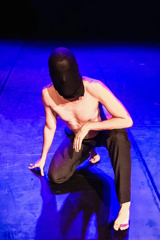 Fotografia de Ricardo Laf mostrando um artista agachado em prontidão no palco, com o rosto coberto, pés descalços e iluminação focada contra um fundo azul intenso no Centro de BH.