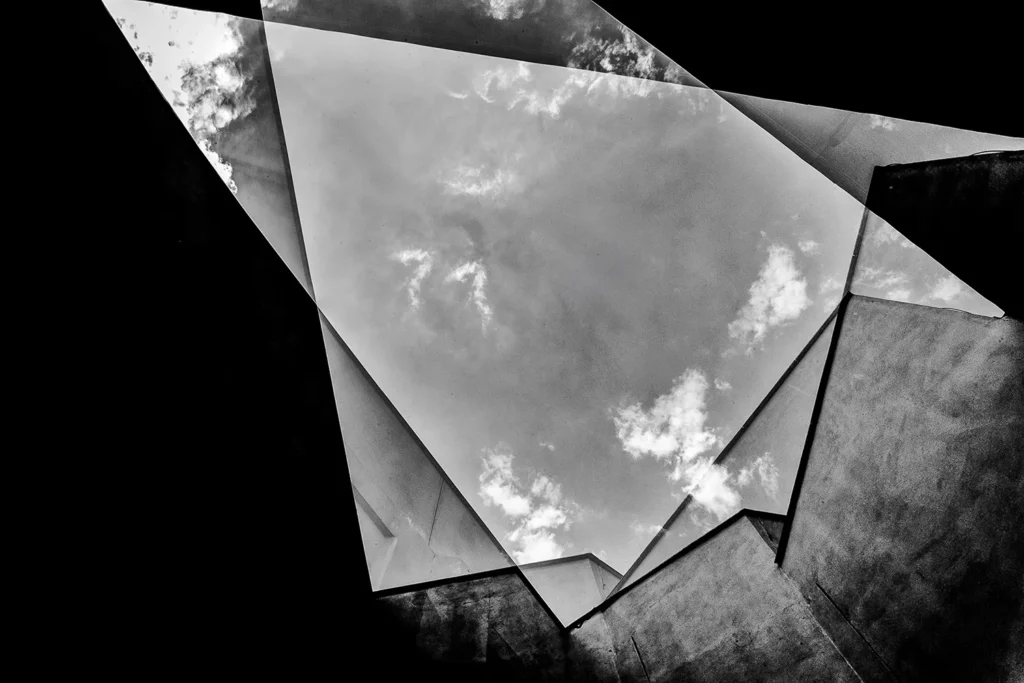 Fotografia em preto e branco com técnica de dupla exposição de Ricardo Laf, sobrepondo planos arquitetônicos geométricos e angulares a uma camada de nuvens densas e texturizadas no céu.