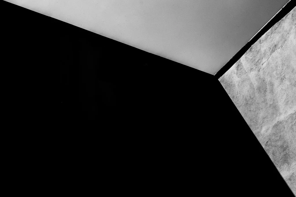 Fotografia em preto e branco de Ricardo Laf capturando uma composição geométrica abstrata, onde uma linha diagonal acentuada divide o quadro entre uma zona de sombra densa e escura e uma parede de concreto com textura áspera e granulada sob iluminação direta.