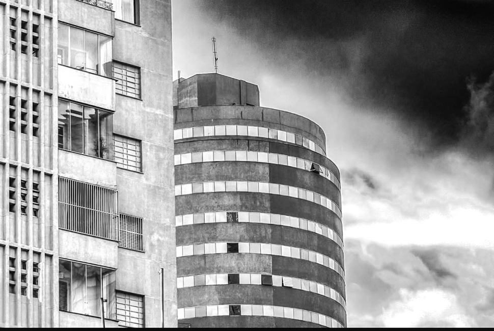 Foto em preto e branco de Ricardo Laf destacando a curvatura e as janelas de um edifício icônico no centro de Belo Horizonte. A perspectiva ressalta a escala monumental da construção contra o céu.