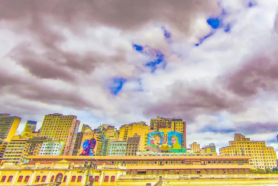 Fotografia de Ricardo Laf no baixo centro de Belo Horizonte. No último plano, edifícios exibem grandes murais de arte urbana em suas paredes, sob um céu com nuvens expressivas.