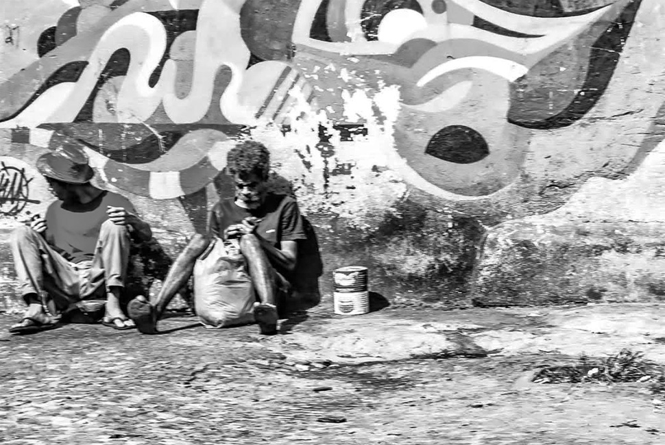 Fotografia em preto e branco de Ricardo Laf feita de dentro de um veículo. A imagem mostra duas pessoas sentadas na calçada no centro de Belo Horizonte, em um momento que sugere a vulnerabilidade do uso de substâncias em ambiente público.