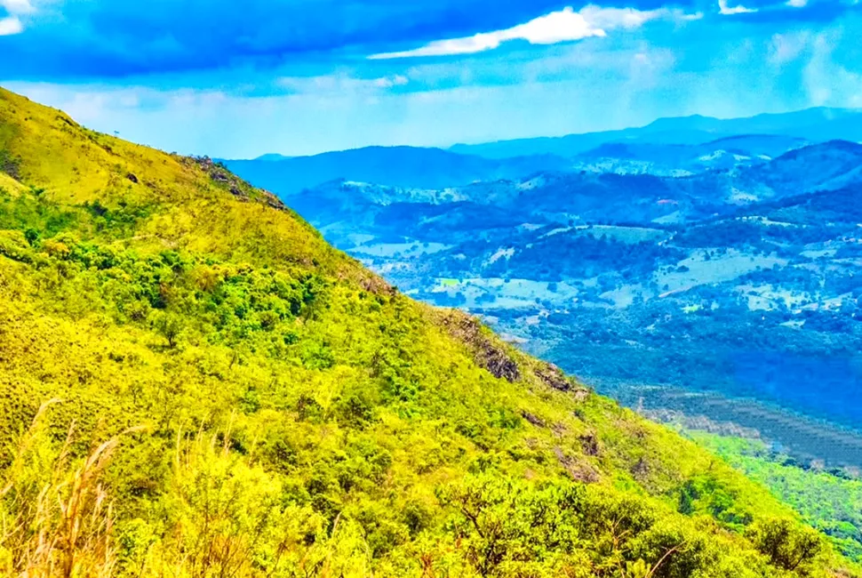 Foto de Ricardo Laf mostrando a vista panorâmica da Serra da Moeda, com encostas verdes em destaque e sucessivas camadas de montanhas azuis sob o céu claro.