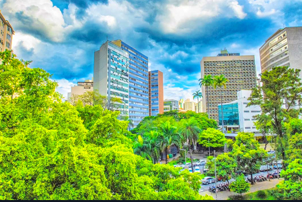 Fotografia de Ricardo Laf mostrando edifícios residenciais e comerciais no centro de Belo Horizonte, cercados por árvores e sob um céu azul, destacando a densidade e a arborização urbana.