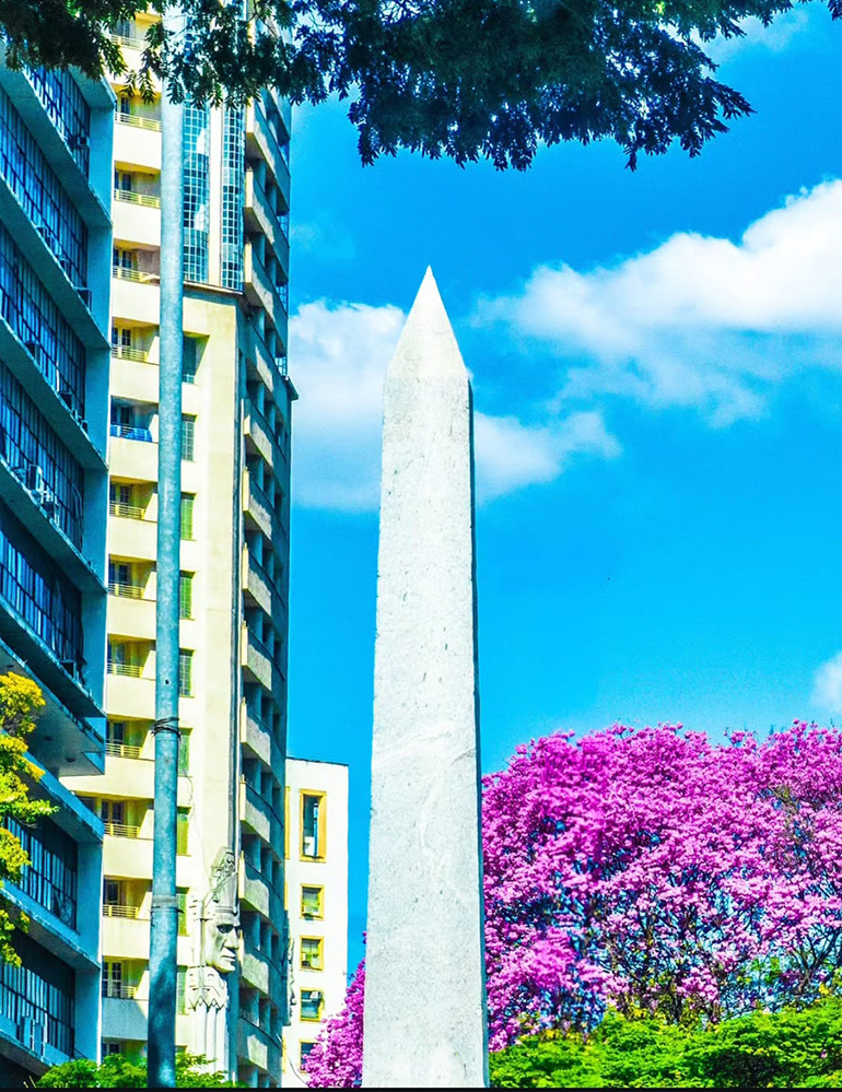 Fotografia de Ricardo Laf do Obelisco da Praça Sete, conhecido como "Pirulito", no hipercentro de Belo Horizonte. O monumento de 1924 é visto em perspectiva vertical contra o céu e cercado por árvores.