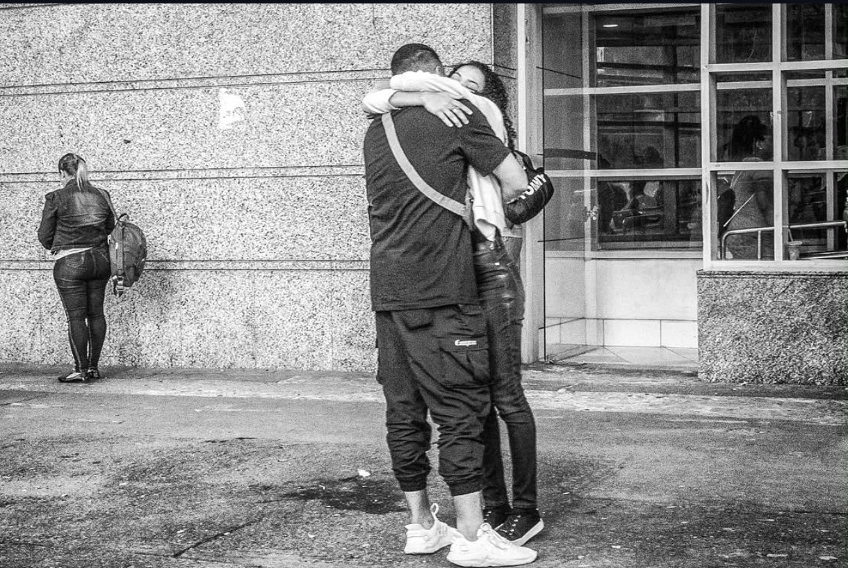 Fotografia em preto e branco de Ricardo Laf no centro de Belo Horizonte. Em primeiro plano, um abraço afetuoso entre duas pessoas; ao fundo, uma mulher de costas voltada para um muro, criando um contraste entre conexão e isolamento urbano.