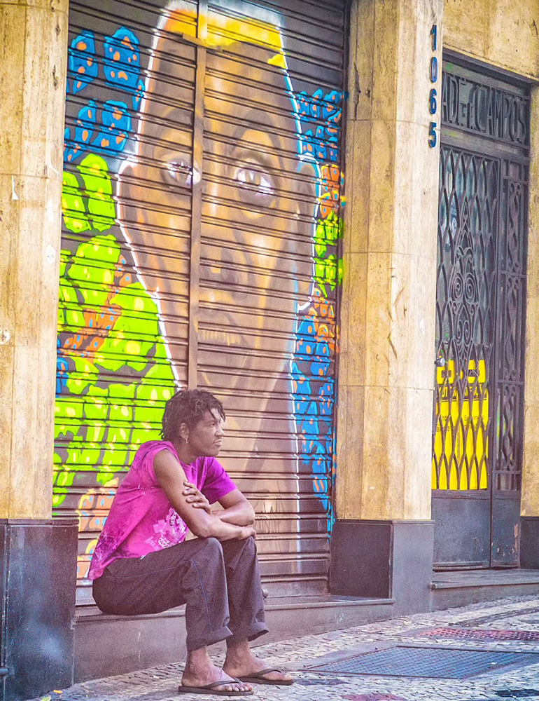 Fotografia de Ricardo Laf feita de dentro de um veículo, registrando um homem sentado na porta de uma loja fechada no centro de Belo Horizonte. O flagrante captura um momento de introspecção e pausa em meio ao cenário urbano.
