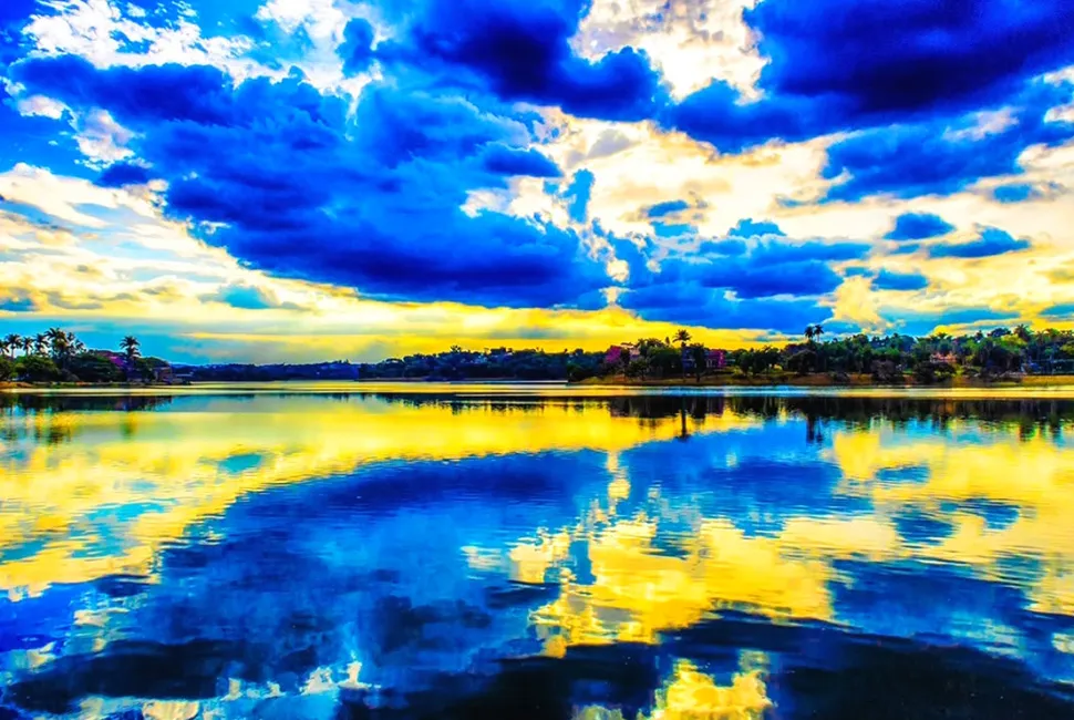 Foto de Ricardo Laf mostrando o reflexo intenso de nuvens brancas e céu azul nas águas da Lagoa da Pampulha, em Belo Horizonte, com vegetação ao fundo.