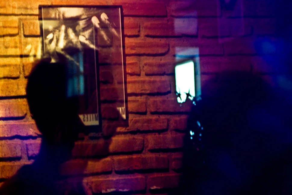 Foto de Ricardo Laf mostrando o interior de um bar em Belo Horizonte à noite, com iluminação suave em tons de azul e roxo e silhuetas de pessoas.