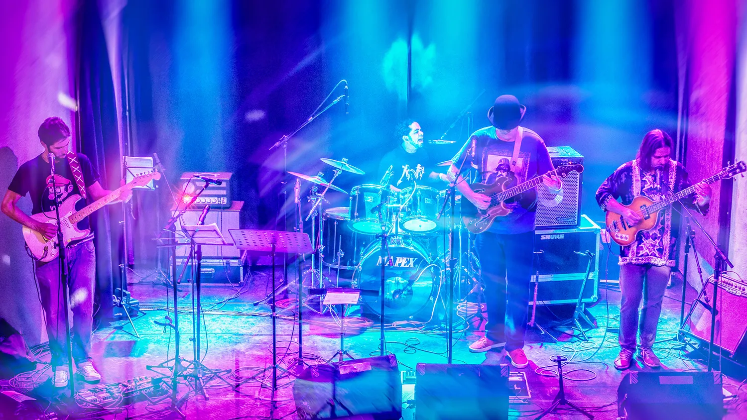 Fotografia de Ricardo Laf apresentando os quatro integrantes da banda Somba em performance coletiva, com iluminação cênica em tons de azul e roxo que destaca a energia do grupo.