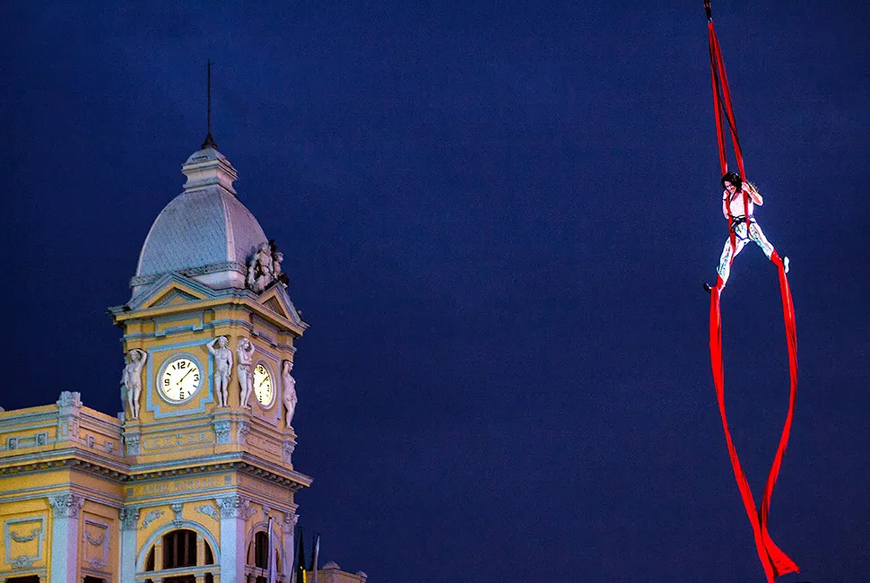 Fotografia de Ricardo Laf mostrando uma performance de tecido acrobático na Praça da Estação durante a Virada Cultural de BH, com um prédio histórico iluminado ao fundo sob o céu noturno.