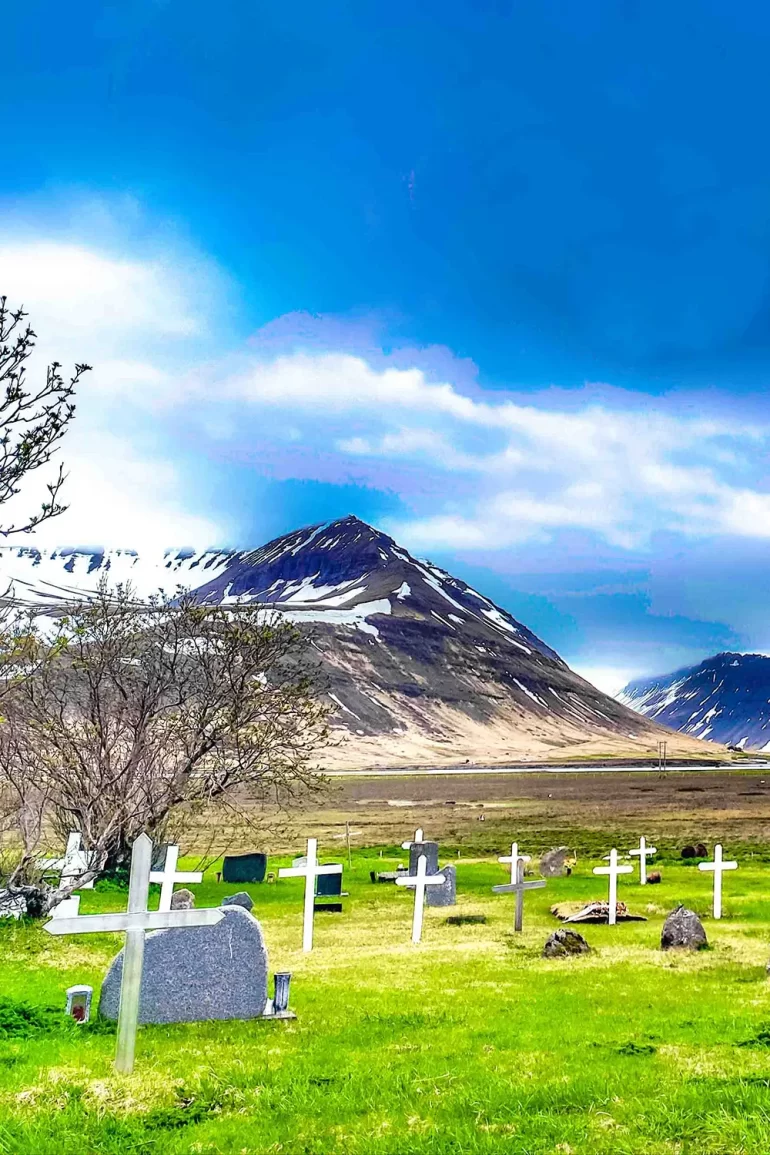 Vista de um pequeno cemitério com cruzes brancas em um campo verdejante, cercado por montanhas nevadas em Holt, Islândia, por Ricardo Laf.