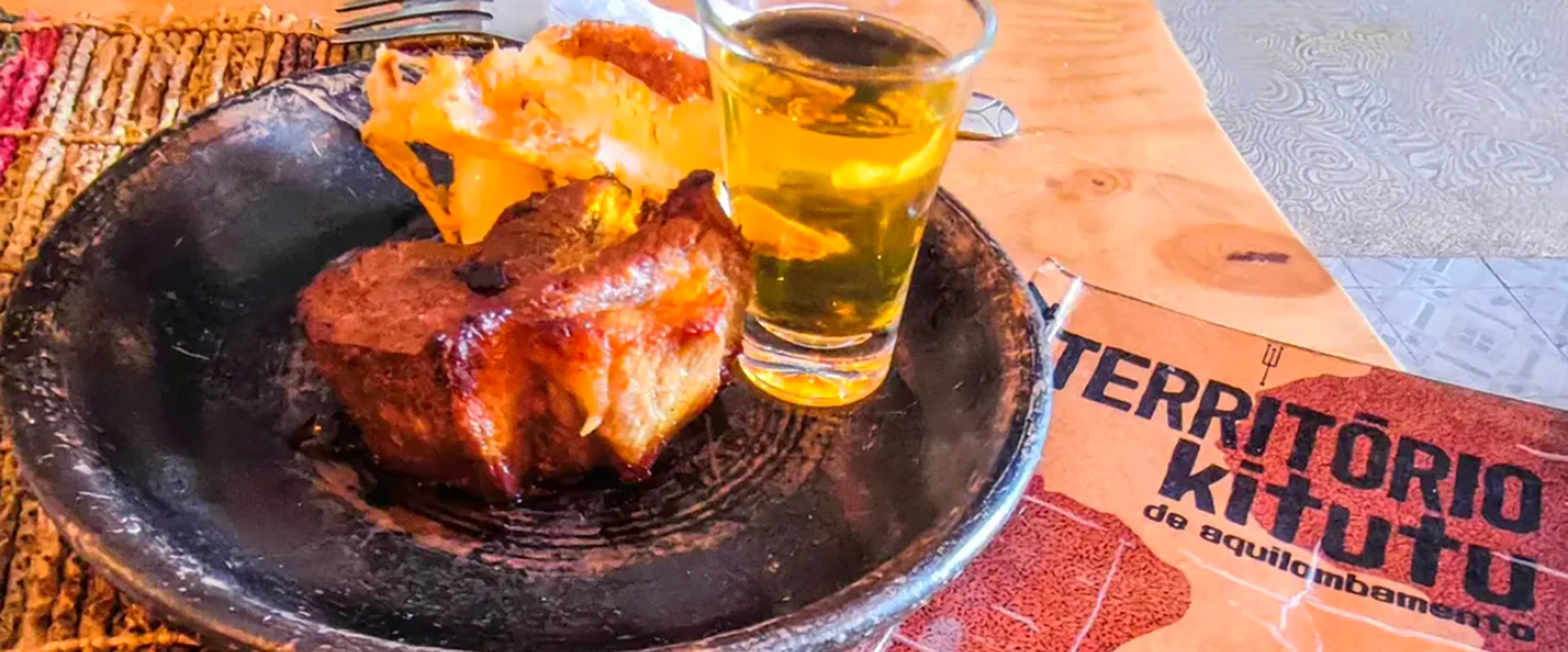 Close lateral de um prato da culinária autoral no Território Kitutu, com um copo de bebida amarela ao lado e o logotipo do local ao fundo.