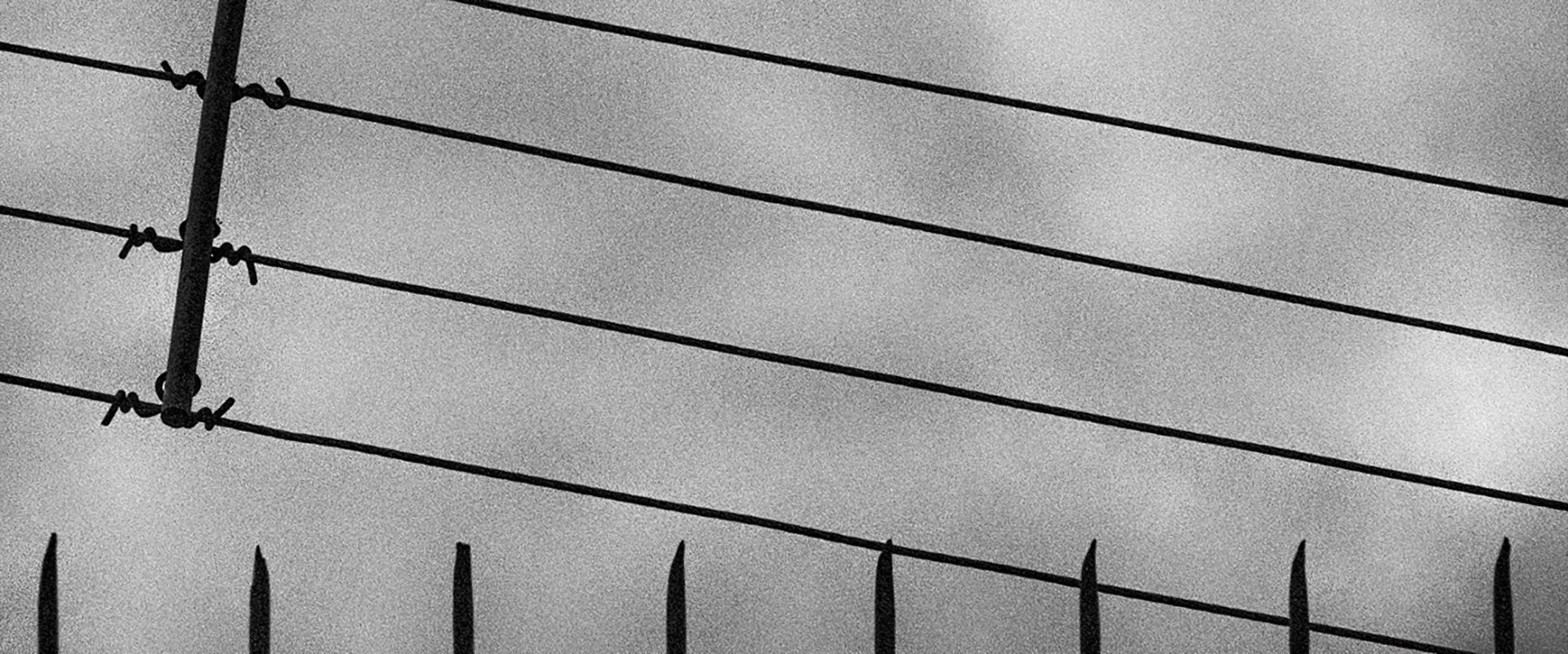 Fotografia em preto e branco com ângulo panorâmico mostrando linhas horizontais de arame farpado sobre um muro, com céu nublado e cinzento ao fundo.