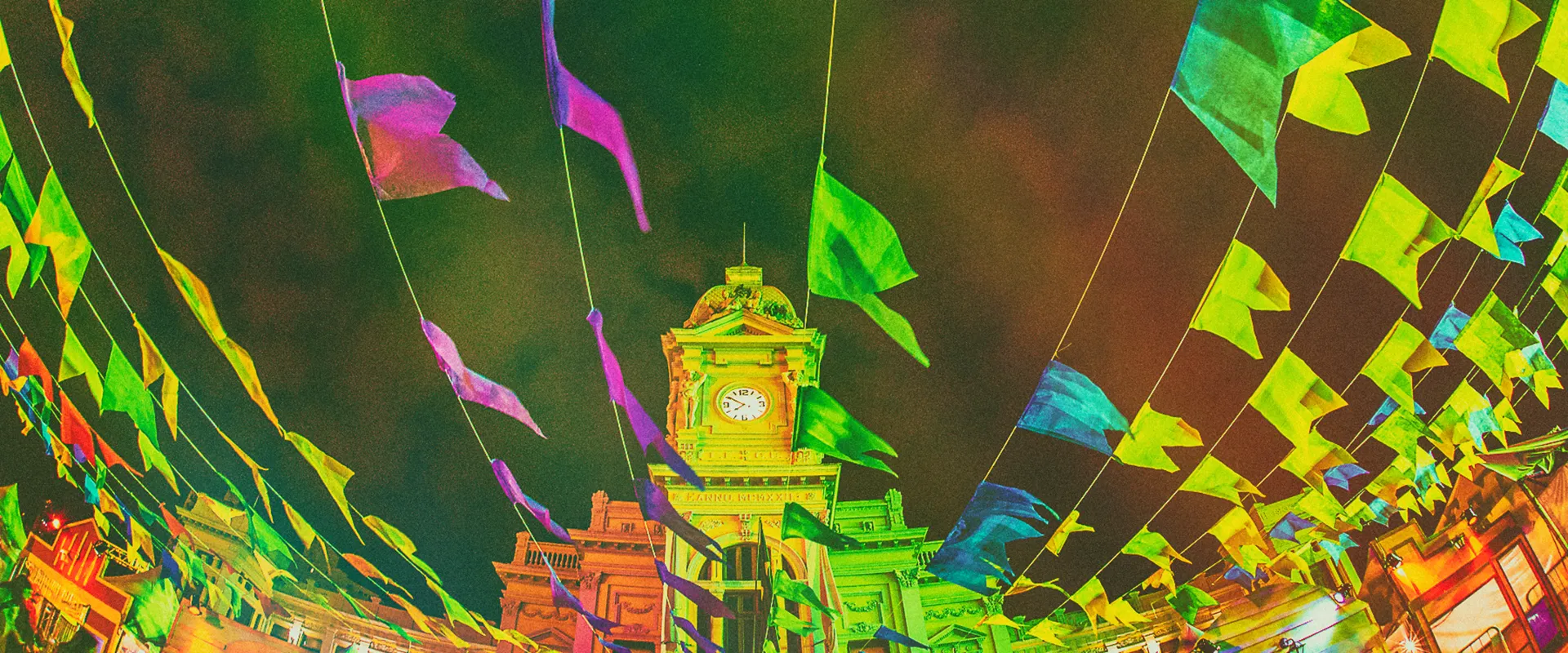 Fotografia de Ricardo Laf mostrando as bandeirinhas coloridas do Arraial de Belô cruzando o céu azul sobre a torre do relógio da Praça da Estação.