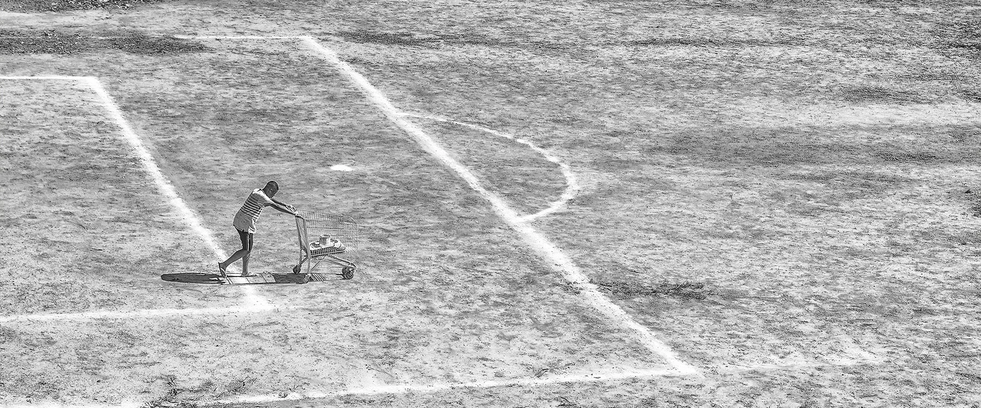 Fotografia aérea em preto e branco capturada por Ricardo Laf mostrando um rapaz de perfil empurrando um carrinho de supermercado de metal pelo chão batido e texturizado do Campo do Inconfidência. Ele caminha sobre a linha branca demarcatória da grande área de futebol. O terreno exibe uma textura seca e granulada.