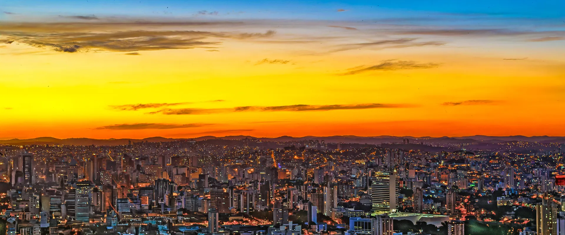 Fotografia panorâmica de Belo Horizonte durante o crepúsculo, exibindo o horizonte urbano iluminado sob um céu em degradê azul e laranja, por Ricardo Laf.