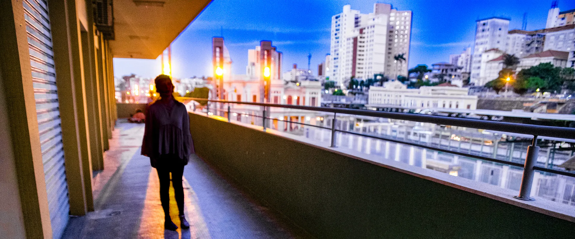 Fotografia colorida de Ricardo Laf mostrando a silhueta escura de uma pessoa de costas, observando a paisagem urbana do Baixo Centro de Belo Horizonte a partir de uma estrutura elevada com guarda-corpo metálico durante o crepúsculo. Ao fundo, edifícios iluminados se destacam sob um céu azul profundo e denso.