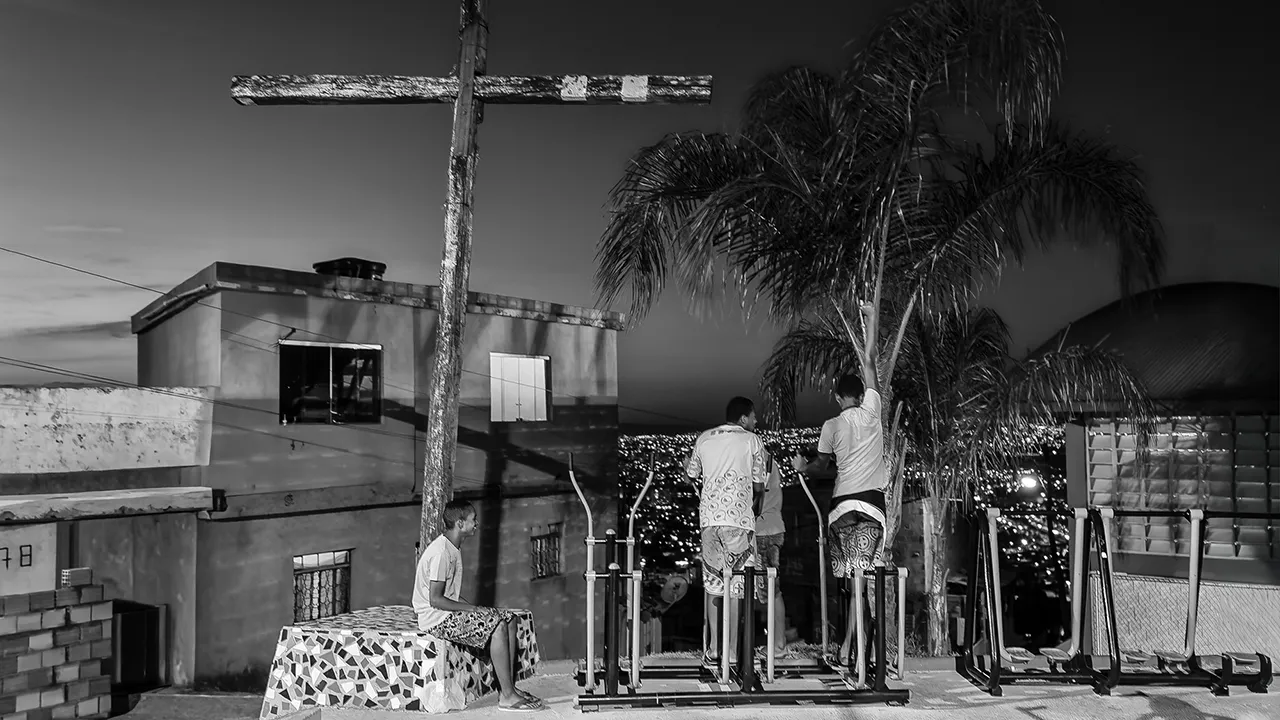 Fotografia em preto e branco de cenário urbano periférico com cruz de madeira e personagens, por Ricardo Laf.