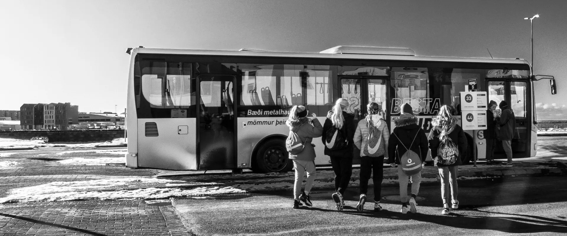 Fotografia urbana em preto e branco de um ônibus de transporte público estacionado em Reykjavik, Islândia. Em primeiro plano, pessoas de costas caminham em direção ao veículo sob um céu com nuvens dispersas. Foto: Ricardo Laf.