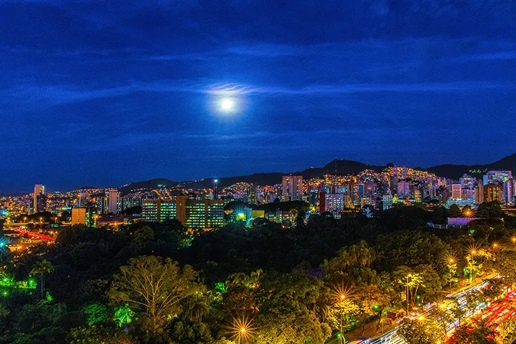 Vista panorâmica aérea e noturna do Hipercentro de Belo Horizonte com rastros de luzes dos veículos e edifícios iluminados sob céu azul-escuro.