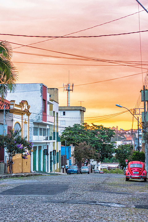 Vista urbana de uma rua na Região Leste de Belo Horizonte com um Fusca vermelho em destaque na paisagem e o horizonte da cidade ao fundo.