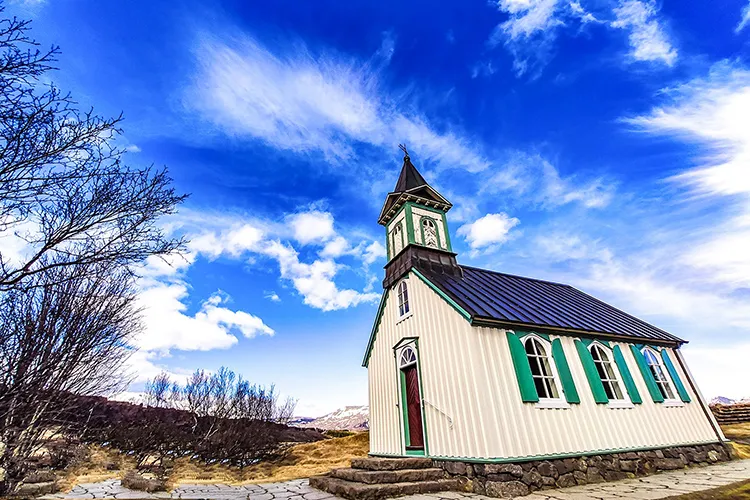 Vista externa da igreja histórica de Þingvallakirkja com teto escuro e campanário de madeira, emoldurada pelas montanhas e pela vegetação do Parque Nacional de Þingvellir, Islândia.