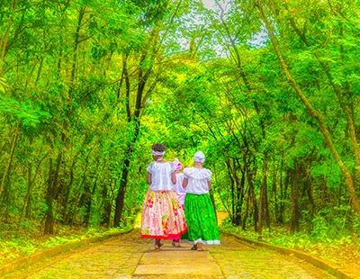 Fotografia em ambiente aberto no Parque Municipal Lagoa do Nado, em Belo Horizonte, com figuras em trajes culturais sob vegetação verde e solar.