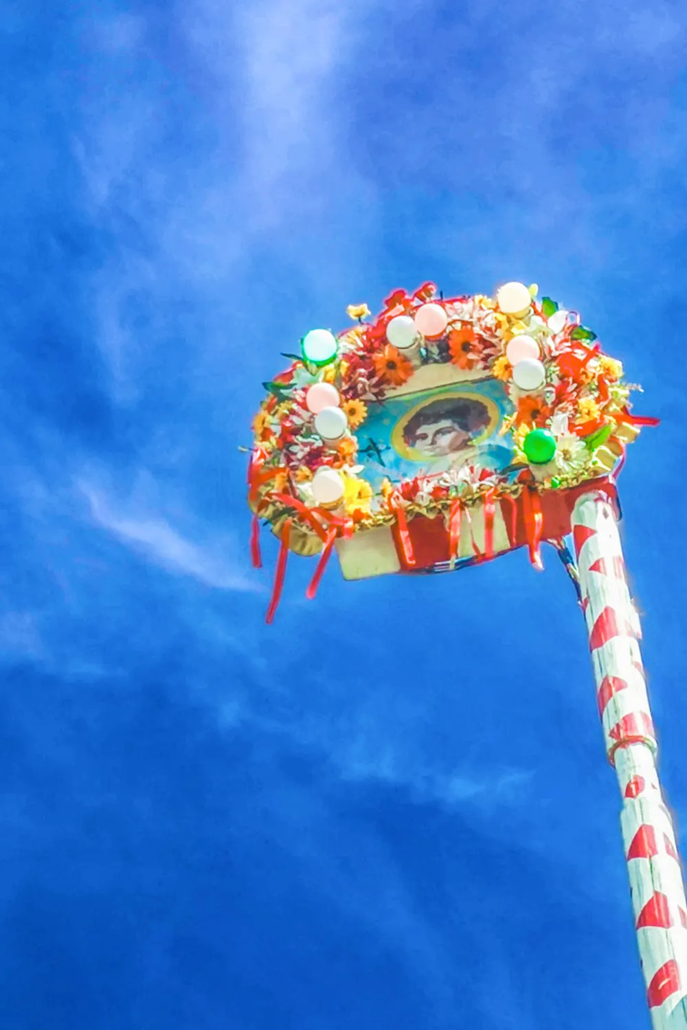 Fotografia em composição vertical de um estandarte festivo de São João, ornado com flores e fitas coloridas, erguido contra um céu azul intenso.