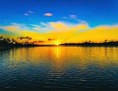 Fotografia do por do sol na Lagoa da Pampulha em Belo Horizonte, com tons alaranjados e amarelos intensos refletidos na água sob céu azul.