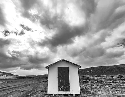 Fotografia em preto e branco de uma pequena estrutura isolada sob um céu dramático com nuvens na região de Kleifaheiði, Islândia.