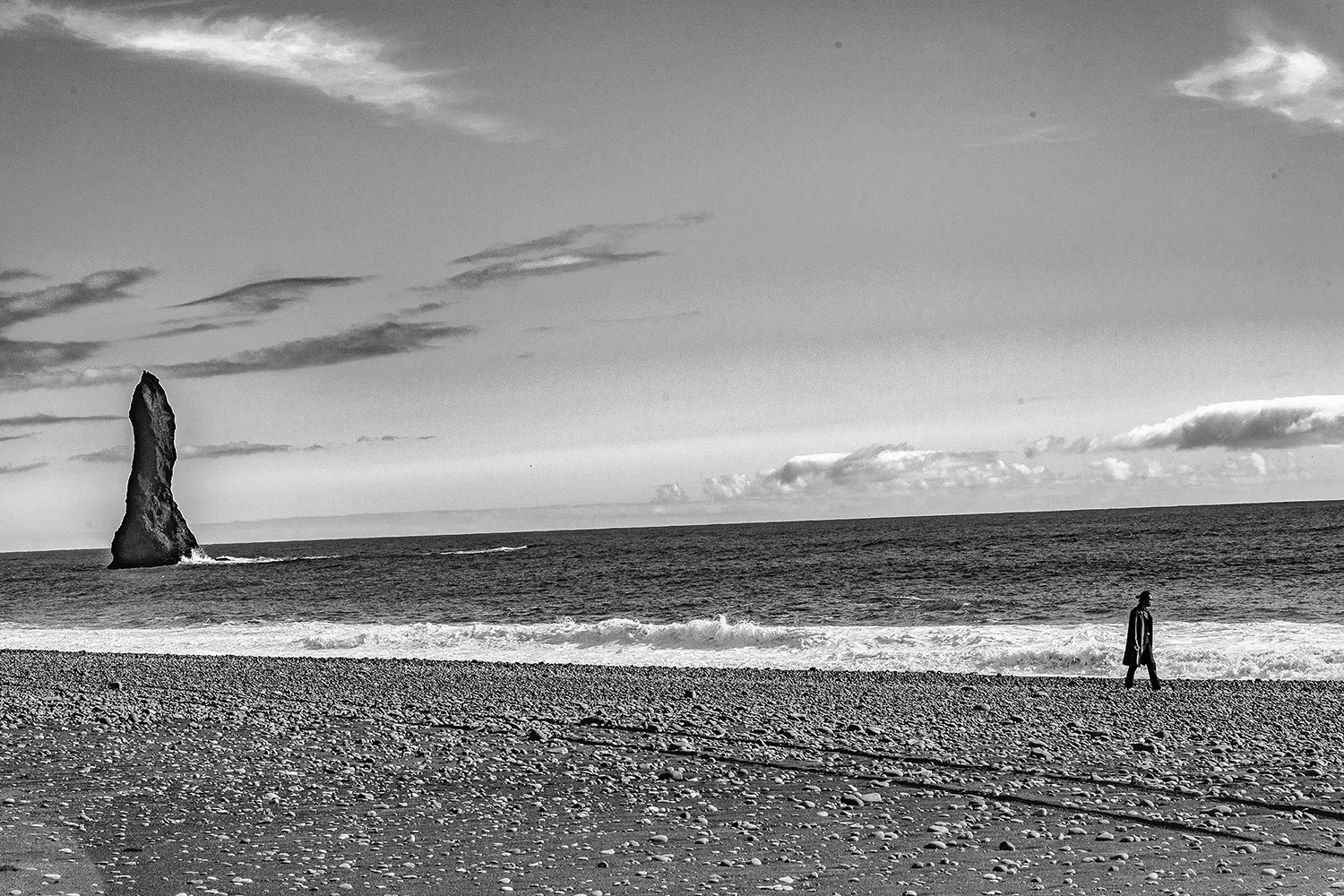 Fotografia em preto e branco de uma praia de areia preta na Islândia, com uma figura solitária caminhando à beira-mar próximo a formações rochosas na água. "The Northern Silence" - Seleção Avançada no Concurso "The Shape of a Moment" | Exposure Photo Gallery