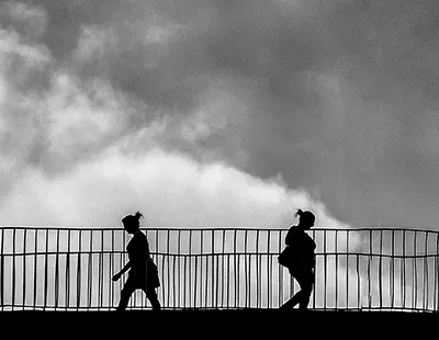Fotografia em preto e branco com silhuetas de pedestres caminhando sobre uma passarela urbana contra céu nublado no Centro de Belo Horizonte.