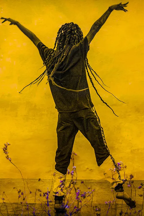 Fotopintura em amarelo intenso com figura corporal em gesto performático sob vegetação e flor roxa em destaque.